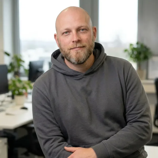 Mann mit Bart im Hoodie sitzt lächelnd in Büro mit Pflanzen.