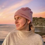 Frau mit Mütze lächelt zum Meer bei Sonnenuntergang, warm bekleidet.
